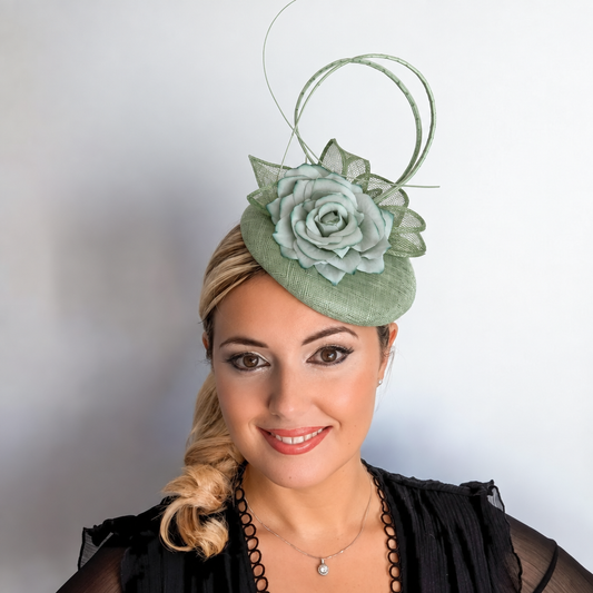 A woman wearing a sage green fascinator hat with a large fabric rose and thin decorative loops, paired with a black top against a plain light gray background.